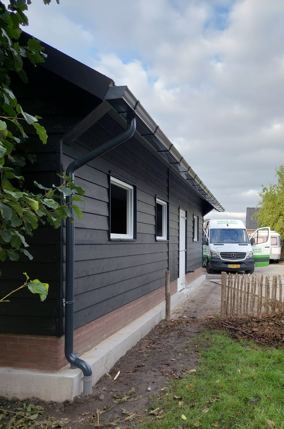 Een foto van onze V150 dakgoot gemonteerd in de kleur antraciet aan een schuur, met daarachter onze montage bus